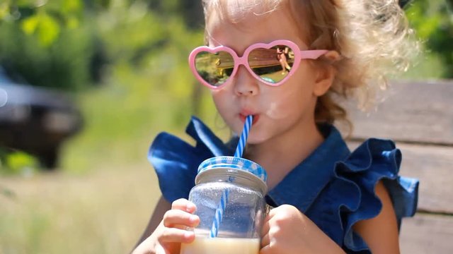 Child girl in sunglasses drinks smoothies in the park on a sunny windy day