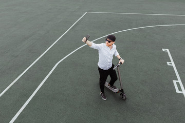 Modern man in stylish outfit making selfie while standing at the street with electric scooter