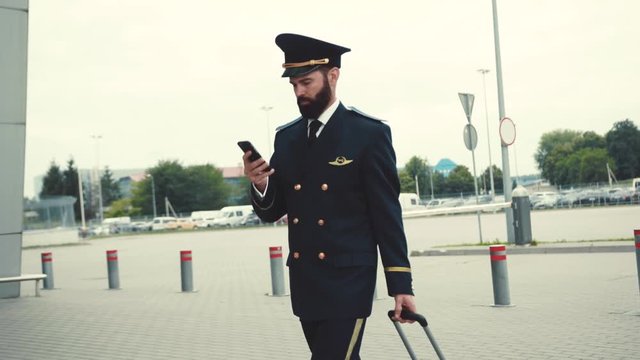 Attractive young caucasian man in pilot uniform pulls his luggage and actively uses smartphone while entering the airport terminal. Doing business, surfing the internet, social networks, being online