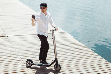 Modern man dressed white shirt and black pants using his phone while standing at a wooden pier with electric scooter