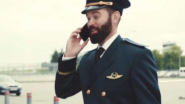 Extremely attractive caucasian bearded pilot in uniform walks by the airport, actively talking on his phone. Modern communication, profession, technologies concept. Male portrait