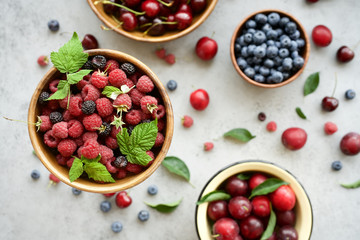 Fresh raspberries, blueberries, plums and cherries in bowls on gray background. Top view
