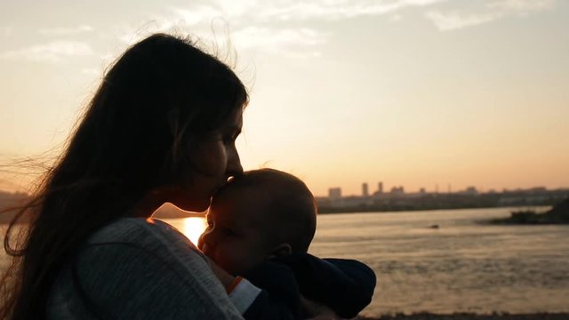 A young mother kisses her little son carefully while standing by the river at sunset.