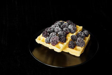 Belgian wafers with frozen blackberries on a black background, close-up.