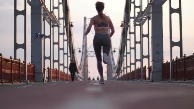 Back view of slim and fit senior redhead woman running across pedestrian city bridge at sunrise. Active attractive female jogging early in the morning on urban steel bridge