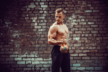 Athletic man working out with a kettlebell in front of brick wall. Strength and motivation. Outdoor workout. Biceps exercise.