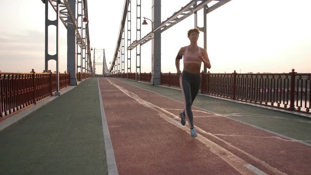 Full length of gorgeous fitness lade in sportswear running on modern pedestrian city bridge at sunset. Healthy active and slim senior woman jogging. Steadicam stabilized shot