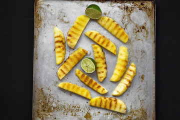 Grilled pineapple with lime on tray over black background, top view.