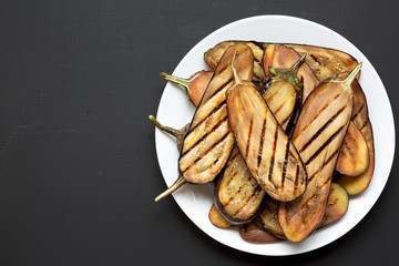 Top view, grilled eggplant on a white plate over black background. Flat lay, overhead. Copy space.
