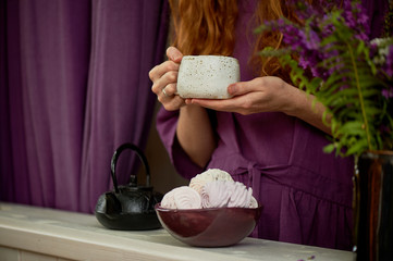 Red-haired girl in a purple dress holding a massive clay mug with tea. Tea on fresh air. Enjoy life