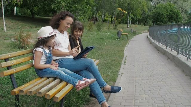 Family with a tablet on the nature. Daughters with mother with a tablet in the park on the bench.