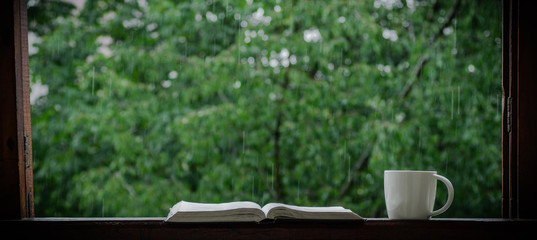 Cozy summer still life: cup of hot coffee and opened book on vintage windowsill and rain outside. Apartment. Rain in the summer