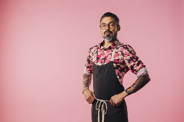 Portrait of a stylish cook with tattoos with hands on waist, isolated on pink studio background