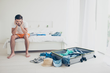 Handsome young bearded confused man wearing white t-shirt, sitting indoors on bed, packing luggage...