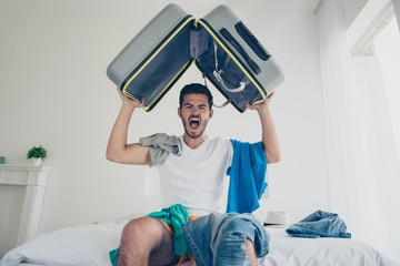 Handsome young bearded brunet evil crazy man wearing white t-shirt, sitting indoors on bed, packing...