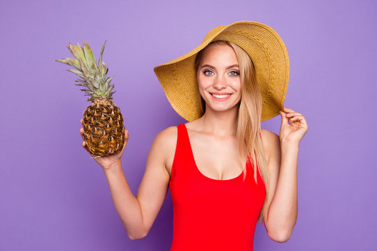 Close up portrait of beautiful and kind girl holds a pineapple in her hand and looks into the camera holding her hat by hand isolated on violet background