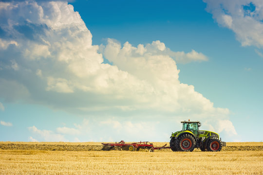 In the summer heat of the tractor plows the field after harvest against the bright Sunny sky with clouds.