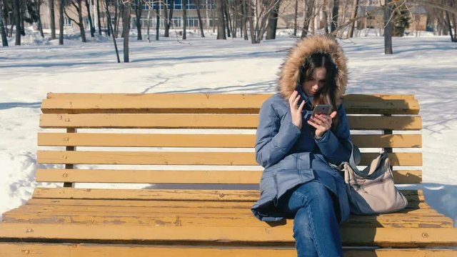 Woman types a message on her phone sitting on the bench in winter city park in sunny day.