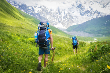 Fototapeta na wymiar Tourists with backpacks on hiking trail walk along green hills in highlands. Hiking in mountains. group of tourists hike to mountain. Leisure. Holidays in mountains. Adventures in Caucasus