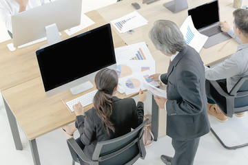 Senior staff in a suit is ordering a female employee to view computerized document at a luxury office desk, with chart and graph on the table.