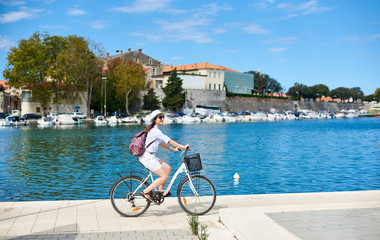 Smiling attractive woman in sunglasses and with backpack riding a bike along stony sidewalk by cozy harbor blue water and resort town cottages on opposite lake shore. Tourism and vacation concept.