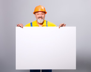 The image of an elderly builder in bright work clothes and a construction helmet that holds a white nameplate for your information