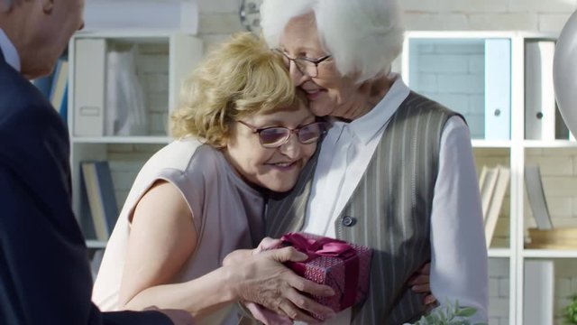 Tilt up of happy elderly businesspeople congratulating retiring female employee packing things into cardboard box: they shaking her hand and giving presents
