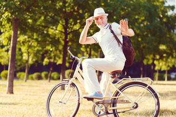 Hey mate. Cheerful elderly gentleman smiling while sitting on his bicycle and waving his hand while welcoming somebody outdoors.