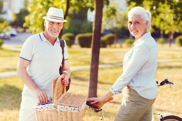 Found a perfect place for our date. Waist up shot of a relaxed retired couple looking into the camera with cheerful smiles on their faces while standing at a bicycle and opening a picnic basket.