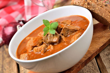 Close-up of pork goulash in a bowl, oregano pepper, onion, red checkered tablecloth in the background