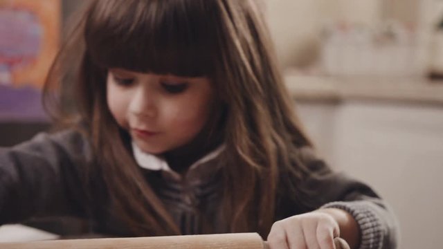 Close up of the cute funny little girl rolling a dough on the wooden table covered with flour with a rolling pin. Portrait shot. Indoor