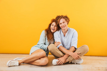 Photo of young happy couple man and woman 20s smiling and hugging while sitting on floor together, isolated over yellow background