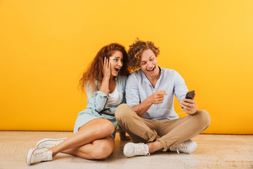 Fototapeta na wymiar Photo of cheerful happy couple man and woman 20s listening to music via earphones while sitting on floor together, isolated over yellow background