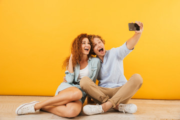 Photo of joyful couple handsome man and curly woman 20s sitting on floor together and taking selfie on smartphone, isolated over yellow background