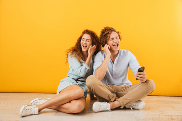 Fototapeta na wymiar Photo of cheerful couple handsome man and curly woman 20s listening to music via earphones while sitting on floor together, isolated over yellow background