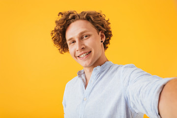 Portrait of curly caucasian guy 20s laughing and taking selfie photo, isolated over yellow background