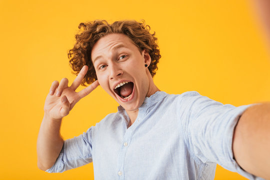Portrait of ecstatic amazed man 20s taking selfie photo and showing peace sign, isolated over yellow background