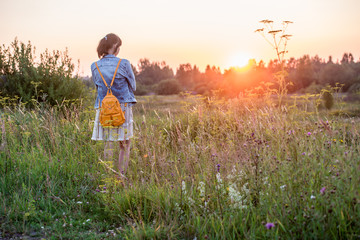 Young girl in a dress and with a backpack in the nature at sunset, rear view