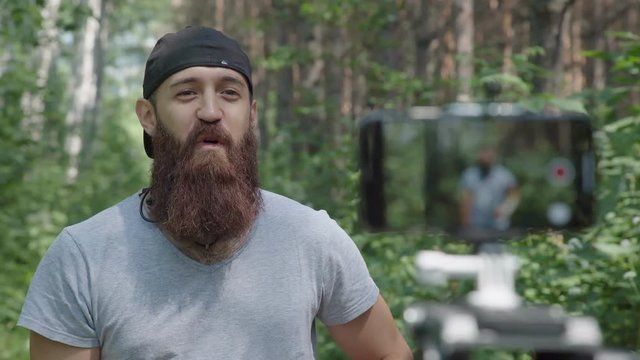 Sporty man in a gray T-shirt and with a beard records a video blog in the forest. Videoblog athlete: hipster does the exercises and shooting them on the camera smartphone. Shooting vlogs, using phone.