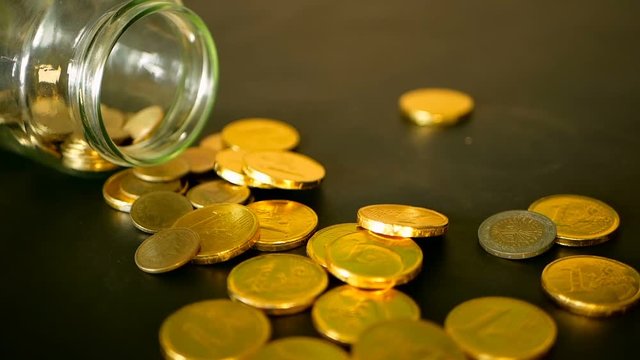 Yellow coins fell out from jar. Symbol of investing, keeping money concept. Collecting cash conis in glass tin as moneybox. Close-up still life with gold coins on black table and rotating penny.