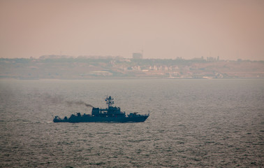 Military ship sailing in the Black sea. A shore with houses is seen at the background. Cloudy foggy weather