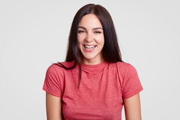 Appealing charming woman with dark hair, healthy skin, smiles joyfully, being in high spirit, wears casual pink t shirt, stands alone against white background. People, happiness and lifestyle concept