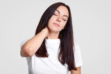 Obraz premium Photo of tired brunette female feels pain in neck as has sedentary lifestyle, needs physical activity, closes eyes, wants sleep, have rest, dressed in casual white t shirt, stands against studio wall