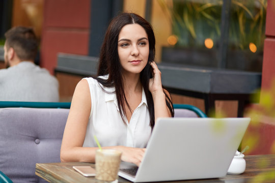 Thoughtful young female in elegant blouse, looks pensively into distnace, works on laptop computer, drinks aromatic coffee, spends free time in cafe outdoor. Woman surrounded with modern devices