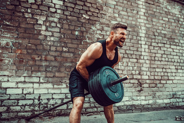 Athletic man working out with a barbell in front of brick wall. Strength and motivation. Outdoor...