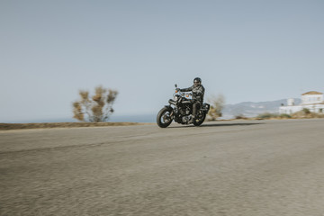 Man in black clothes riding an American motorcycle fast in a road in the mountain, with blue sky in the background.