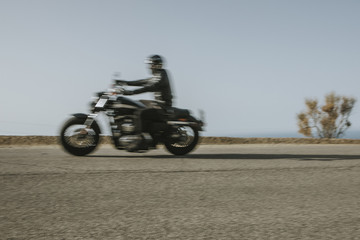 Man in black clothes riding an American motorcycle fast in a road in the mountain, with blue sky in the background.