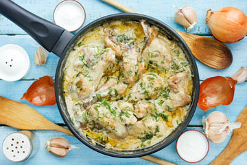 Gedlibzhe — kabardian chicken in sour cream sauce with onion served in black frying pan on a blue wooden background with ingredients top view