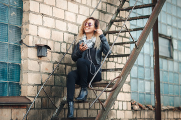 Smiling young woman in mirrored sunglasses, a black leather jacket, black jeans sitting on an urban metal stair against a brick wall. Woman listening to music on the stairs of industrial building