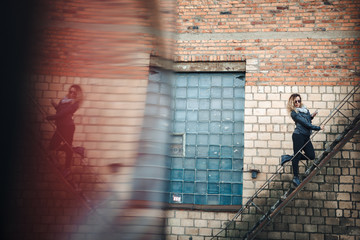 Smiling young woman in sunglasses, a black leather jacket, black jeans standing on an urban metal stair against a brick wall and dances. Woman listening to music on the stairs of industrial building.